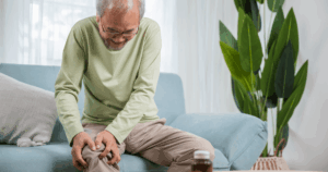 Old man sitting on a couch, clutching his knee in pain.